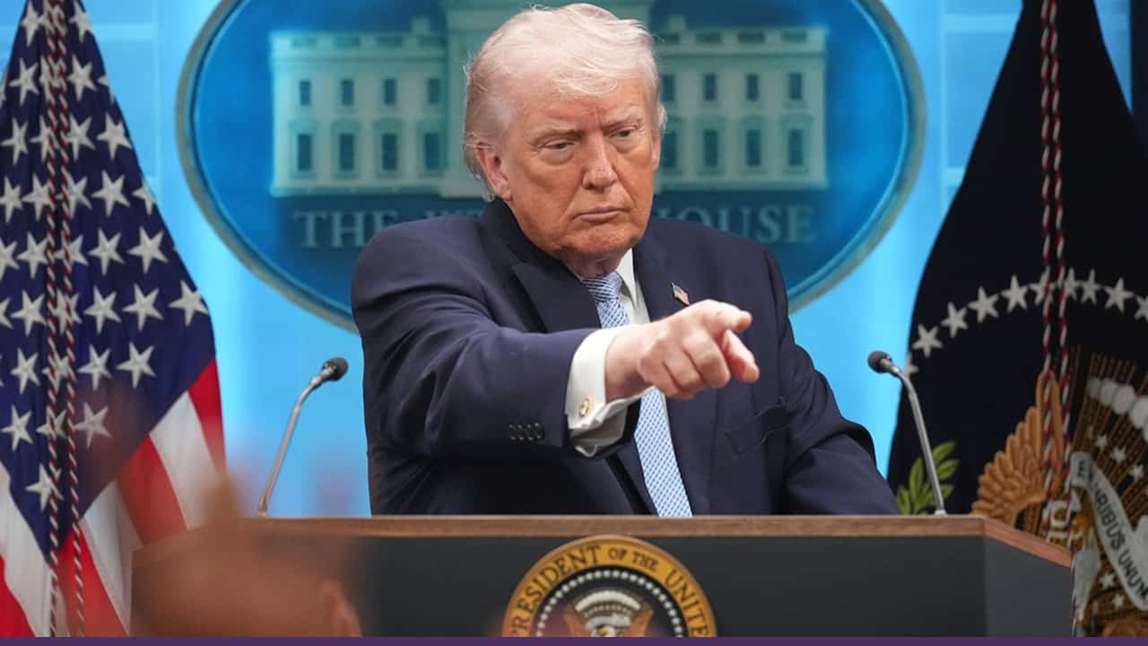 A man at a White House press briefing points toward someone while standing at a podium, with U.S. flags in the background.