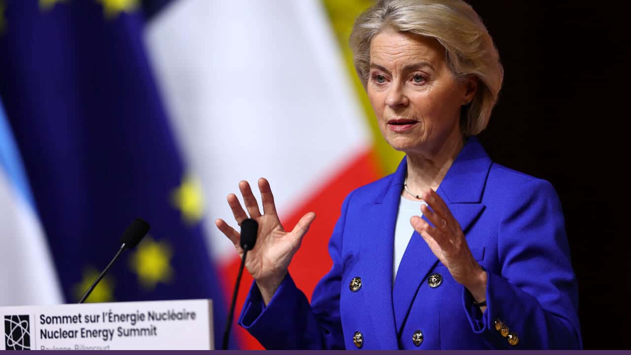 Ursula von der Leyen speaking at a podium during the Nuclear Energy Summit, gesturing with both hands, with EU and national flags blurred in the background.