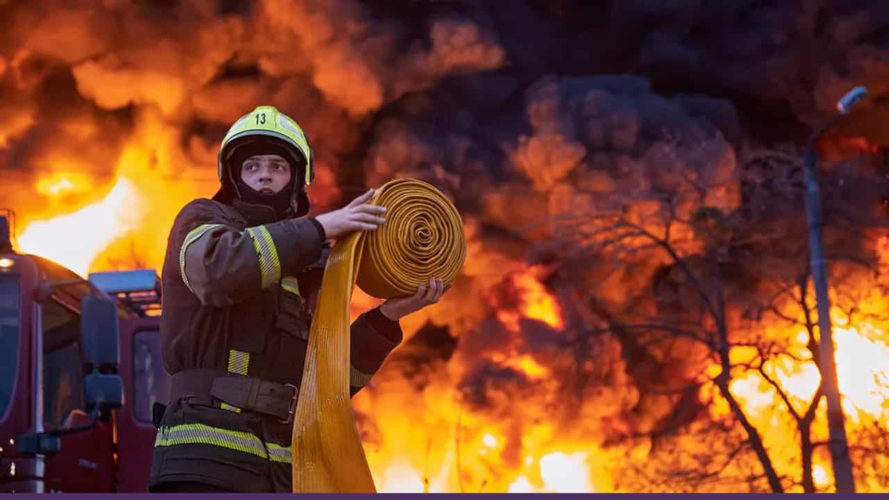 A firefighter in protective gear holds a coiled fire hose while standing in front of a massive blaze, with thick black smoke and towering flames engulfing trees and structures in the background.