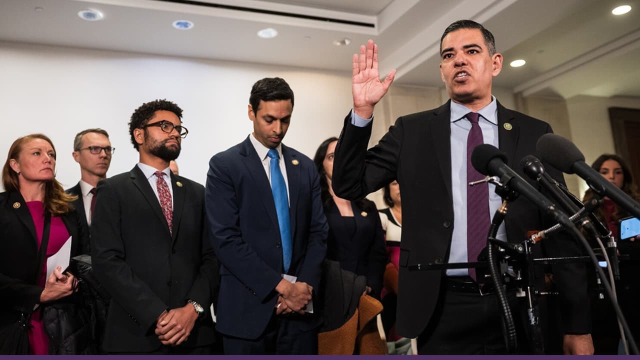A group of U.S. lawmakers stand at a press conference, with one speaker raising his hand while addressing reporters at microphones, as colleagues stand behind him listening attentively.