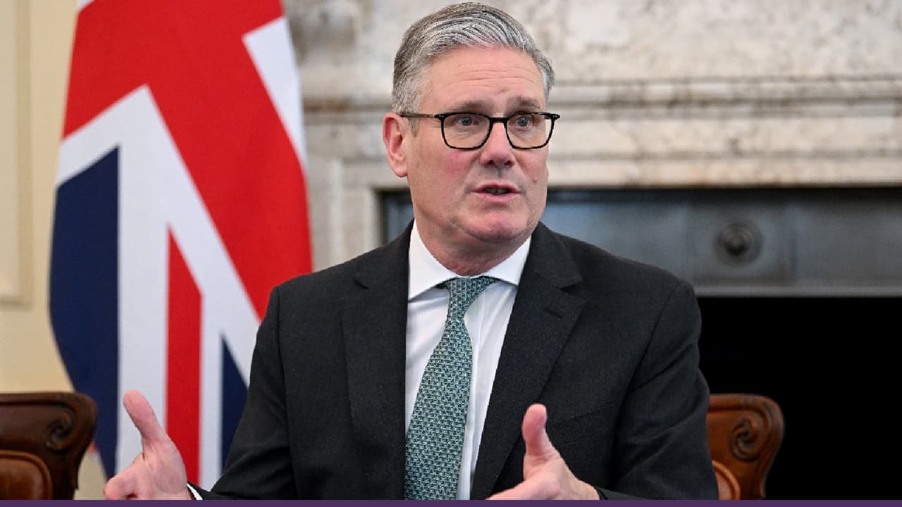 A man in a suit and glasses speaks while gesturing with his hands, seated indoors with a Union Jack flag and a fireplace in the background.