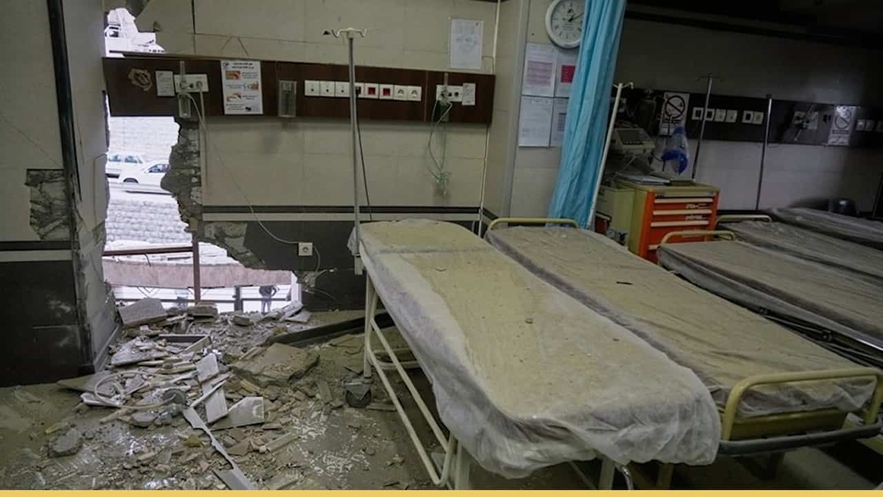 A damaged hospital room with two empty beds covered in dust, debris scattered across the floor, and a large hole in the wall exposing the outside.