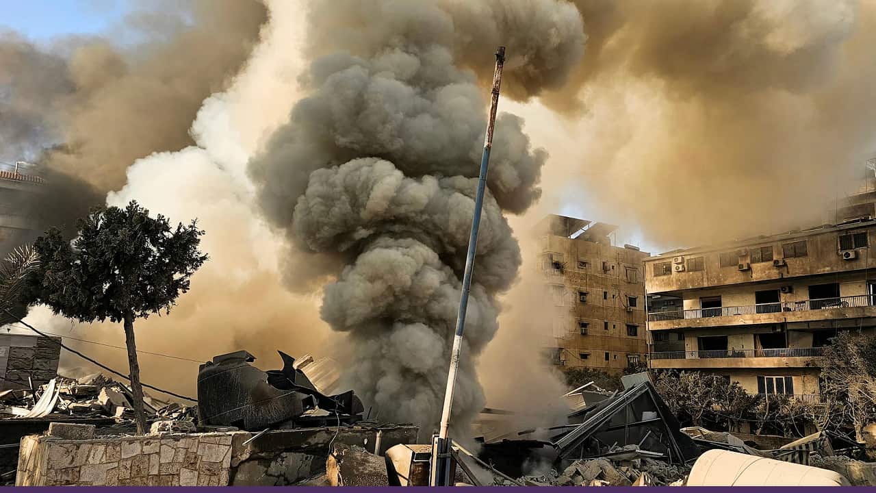 Thick plumes of smoke rise from heavily damaged buildings and debris following an apparent airstrike in an urban area.