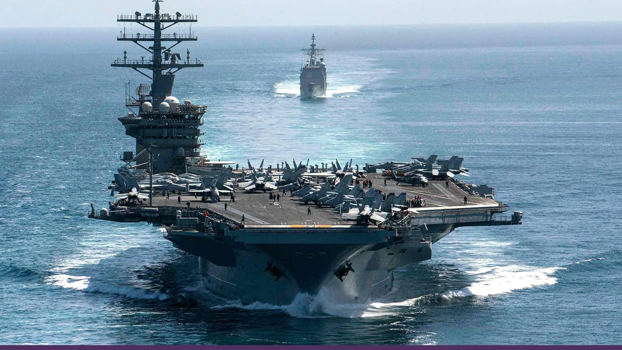 US aircraft carrier sailing in open waters with fighter jets on deck and a destroyer following behind