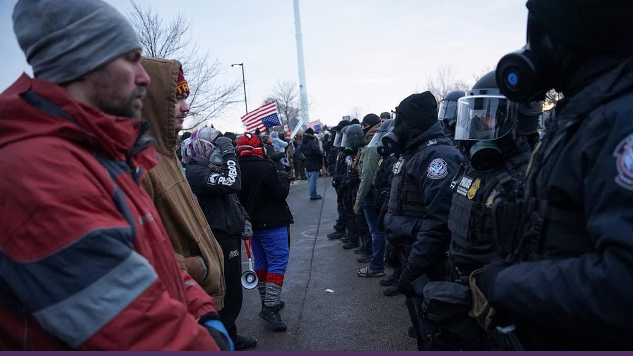 Protesters stand face-to-face with a line of heavily equipped police in winter weather, with US flags visible in the background