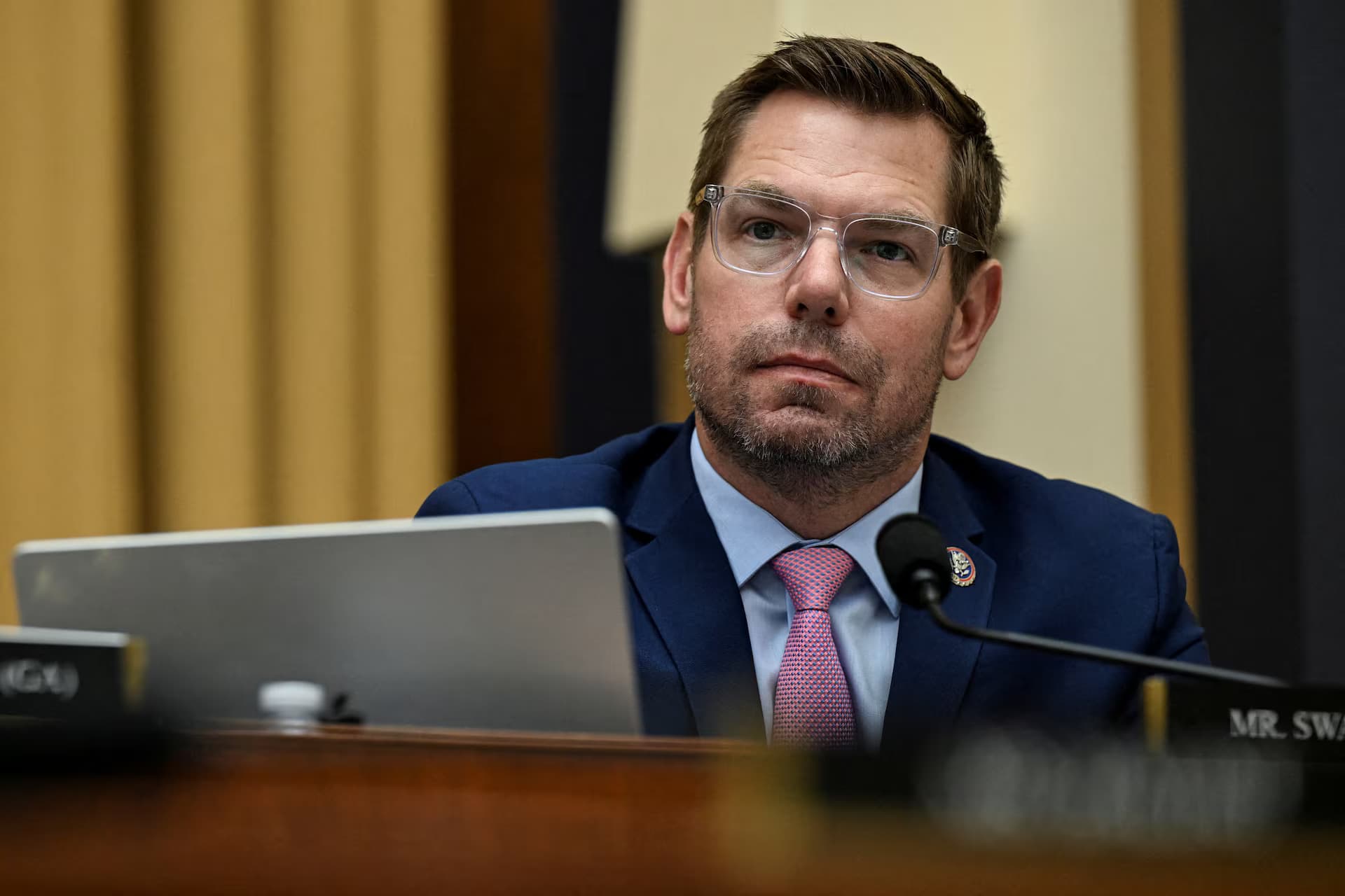 A man in a suit and glasses sits at a desk with a microphone and laptop, appearing to listen attentively during a formal hearing or meeting in a government setting.